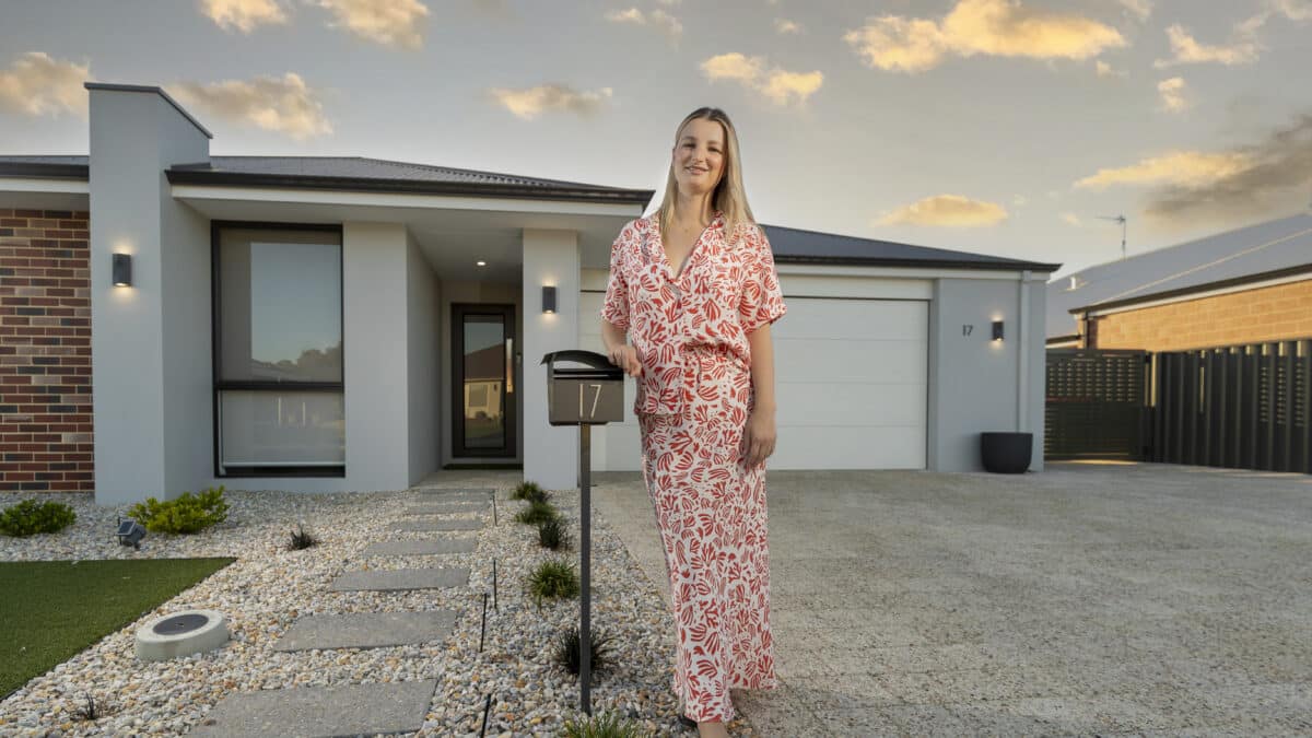 First home buyer stands at the front of her modern house leaning on her mailbox. There are concrete steps with pebbled landscape walking up to the front door. A modern home with dark trim window frames, glass door and red bricks are featured on the facade.
