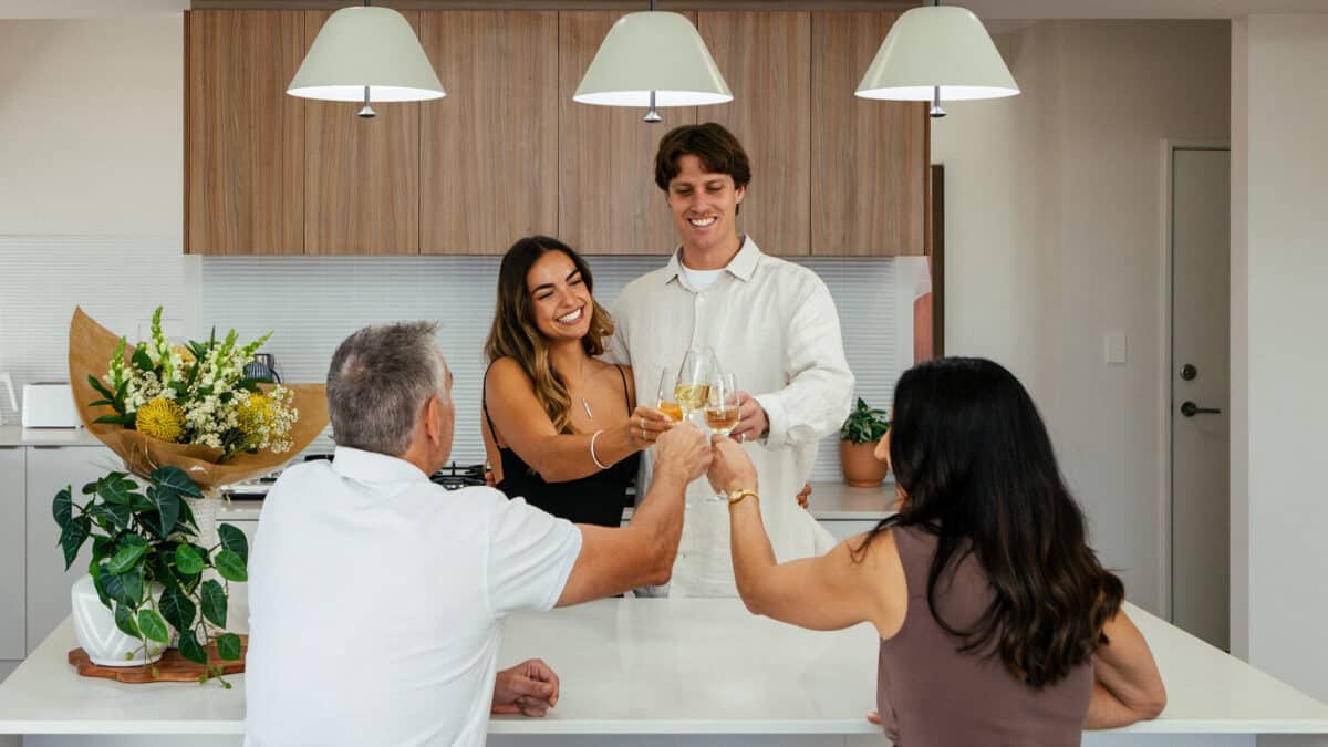Parents congratulate with cheers of bubbles at the kitchen island bench with first home buyer couple in their first home. This Step 1 Homes home design features premium lighting fixtures, horizontal kitkat tiles for the kitchen splashback, waterfall edge kitchen island, and a neutral colour palette.