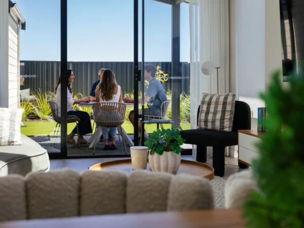 First home buyer, her partner, and parents sit outside in the outdoor entertaining alfresco area. The home design features an open-plan living space that opens out to the shaded alfresco area, where her partner and parents sit. Lush green lawns extend further into the backyard, perfect for their dog.