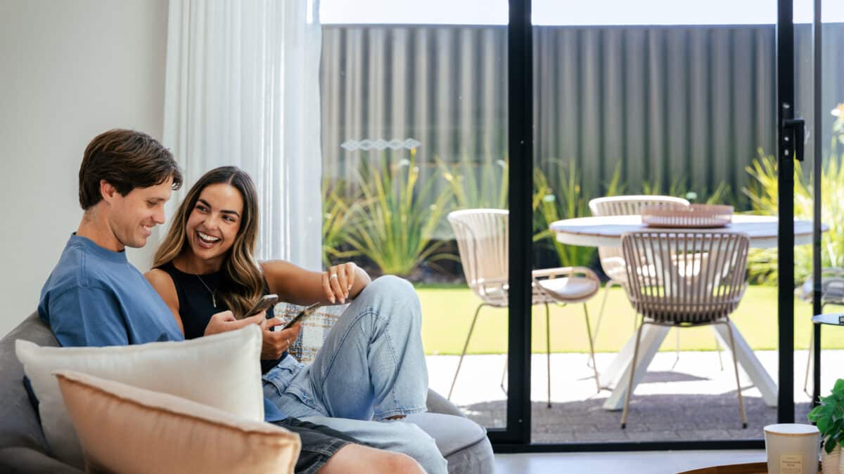 First home buyer couple sit together on the couch with their phones out. The home design shows an open plan living area that opens up to the outdoor alfresco area with large glass panel doors. The background shows a sunny day with a shaded outdoor dining area, and lush green lawn and green landscaping.