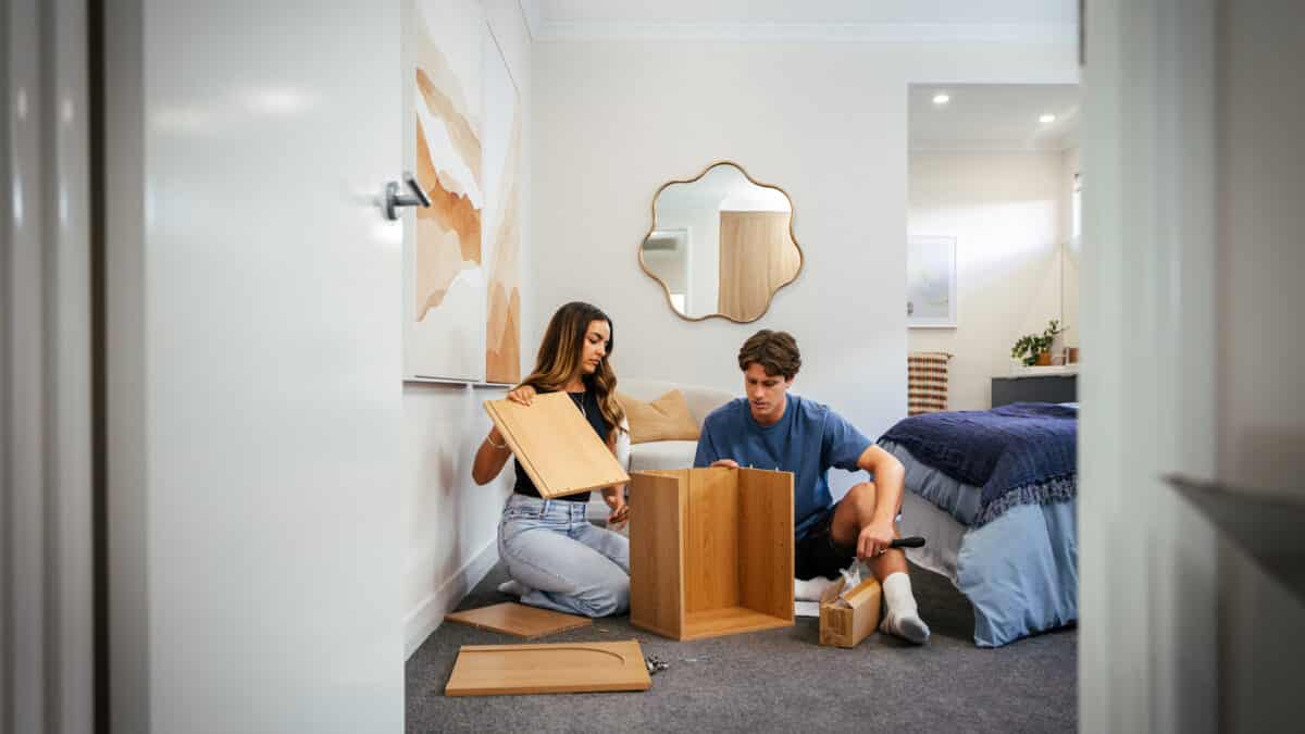 First home buyer couple work together to put a piece of furniture together in their master bedroom. The home design shows a master bedroom with dark coloured carpet, modern downlights, and an open ensuite from the master bedroom.
