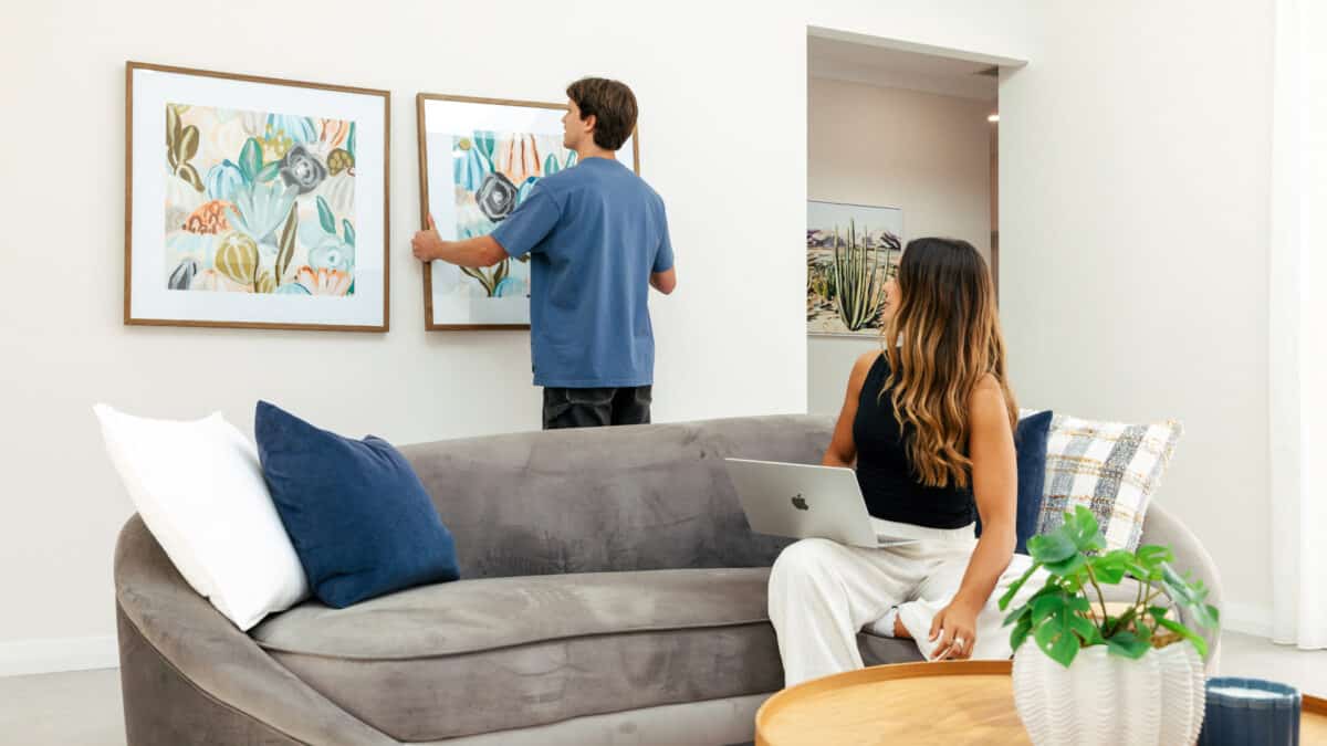 The modern home design for first home buyers built by Step 1 Homes. First home buyer sits on the couch in the lounge room with a MacBook Laptop, while her partner puts up a picture frame on the wall.