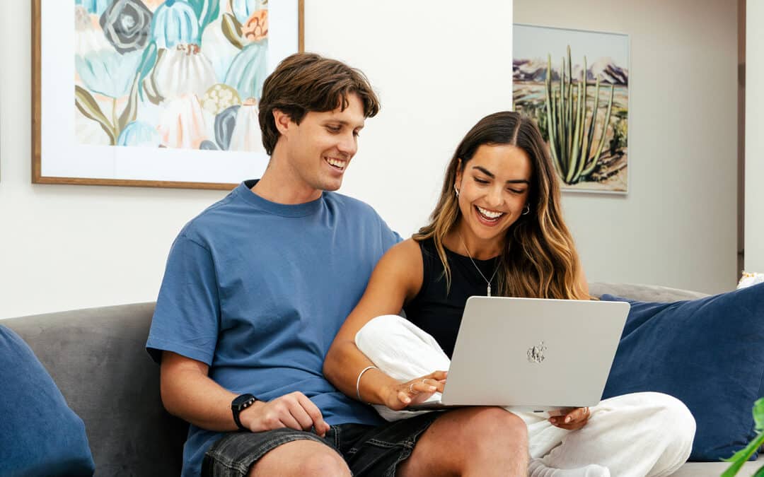 The modern home design for first home buyers built by Step 1 Homes. First home buyer sits on the couch in the lounge room with a MacBook Laptop with her partner.