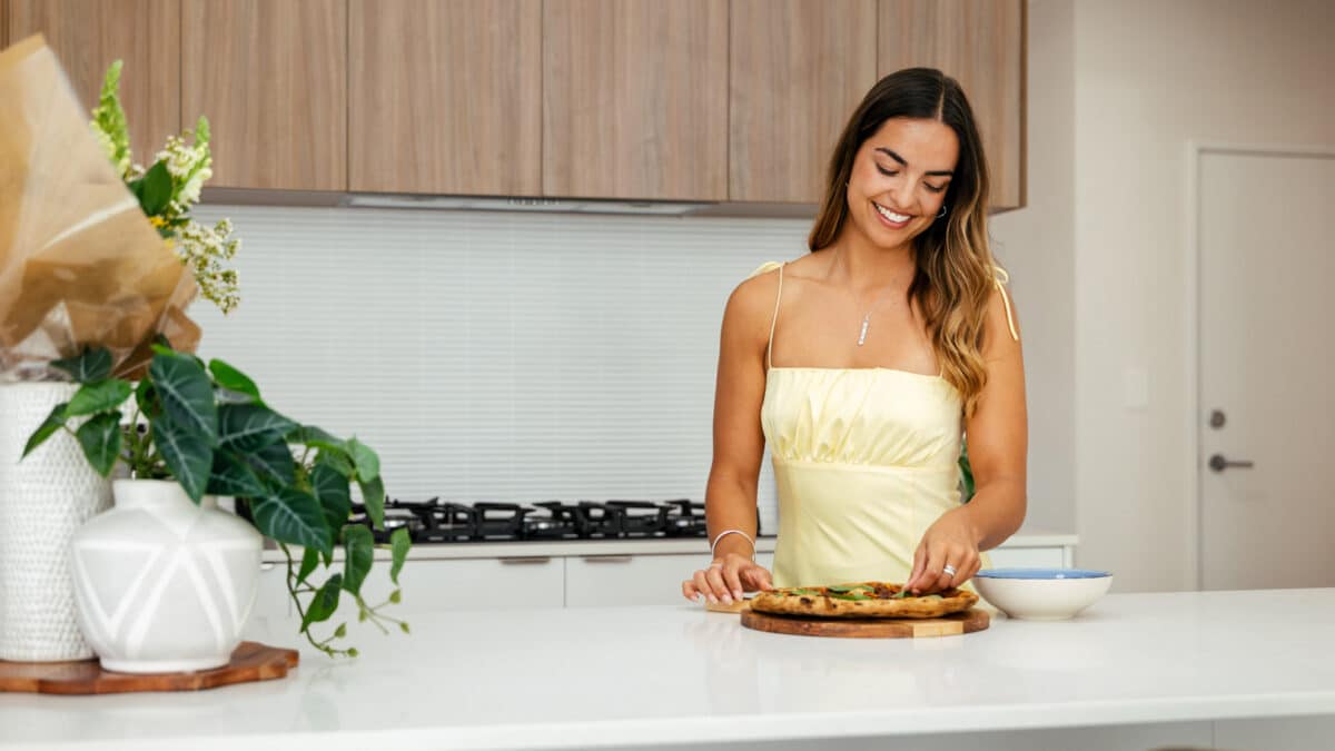 First home buyer in her kitchen island bench putting basil on top of the pizza. The home design features a white stone benchtop, contrasted by warm wooden cabinetry in the kitchen. Built by Step 1 Homes.