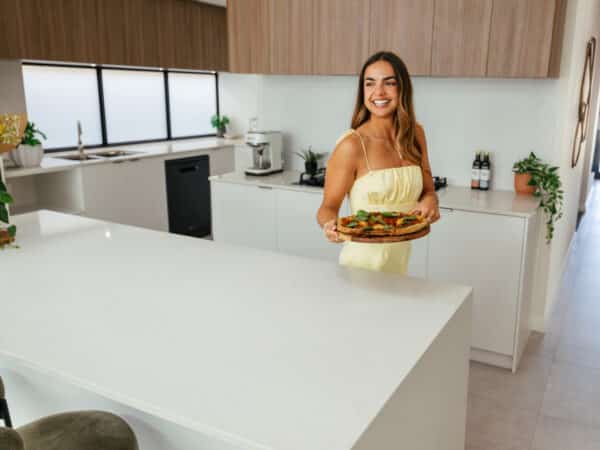 First home buyer in her kitchen island bench brings pizza out for guests. The home design features a white stone benchtop, contrasted by warm wooden cabinetry, and horizontal kitkat tiles in the kitchen. Built by Step 1 Homes.