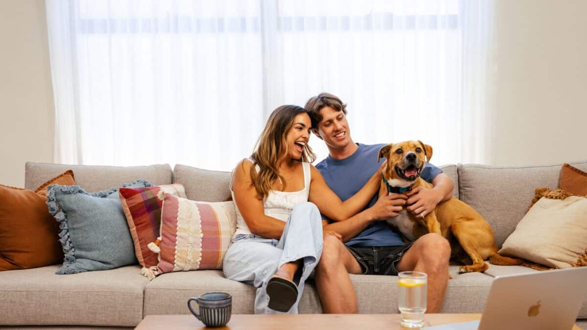 First home buyers sit in their living room on their couch of their first home, with their dog who has joined them on the couch. New generation of home design and home ownership focuses on smart and practical living, including the use of large windows to let in lots of natural light.