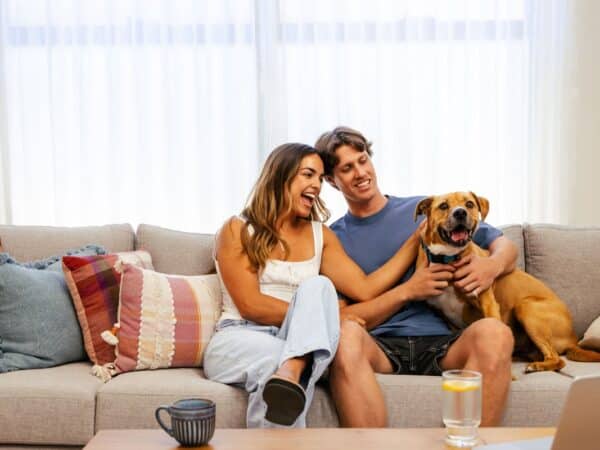First home buyers sit in their living room on their couch of their first home, with their dog who has joined them on the couch. New generation of home design and home ownership focuses on smart and practical living, including the use of large windows to let in lots of natural light.