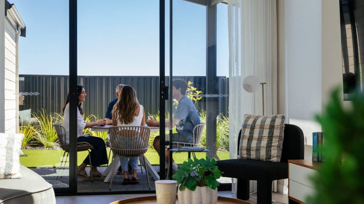 First home buyer, her partner, and parents sit outside in the outdoor entertaining alfresco area. This Step 1 Homes home design features an open-plan living space that opens out to the shaded alfresco area, where her partner and parents sit. Lush green lawns extend further into the backyard, perfect for their dog.